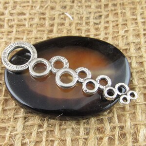 May include: A silver-toned metal toggle clasp component featuring a series of connected circular rings of varying sizes, with the largest ring at one end displaying a textured, grooved surface pattern along its outer edge.