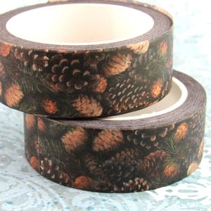 Könnte beinhalten: Zwei Rollen dekoratives Washi-Tape mit einem Waldmuster aus Tannenzapfen und Nadelbaumzweigen. Das Klebeband hat einen dunkelbraunen Hintergrund mit orangefarbenen und grünen Akzenten, ideal für Bastelarbeiten und Scrapbooking.