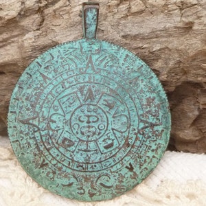 Puede incluir: Un colgante redondo de color turquesa con un diseño en relieve detallado. El colgante presenta una figura central en forma de sol rodeada de intrincados patrones y símbolos. Tiene un lazo en la parte superior para colgar y una apariencia envejecida y antigua.