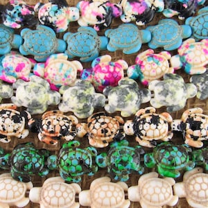 May include: A collection of turtle-shaped beads arranged in horizontal rows. The beads feature various colors and patterns, including solid turquoise, speckled black and white, vibrant pink and blue splatters, and solid cream, all with detailed shell textures.