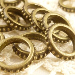 May include: A close-up of a pile of antique gold-toned metal jump rings. The rings are round and have a beaded edge.