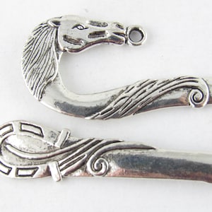 Puede incluir: Dos abalorios de marcapáginas de metal plateado. Un abalorio presenta un diseño de cabeza de caballo con una cola rizada. El otro abalorio presenta un diseño de herradura con una cola rizada.