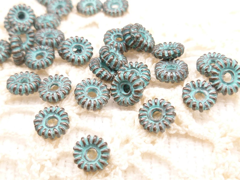 May include: A collection of turquoise-colored, flower-shaped beads with a distressed, antique finish. Each bead has a central hole and a detailed, ridged edge. The beads are scattered on a cream-colored, textured surface.