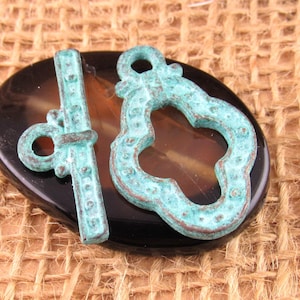 Può includere: Un set di chiusura a T composto da una barra e un anello a forma di quadrifoglio. Entrambi i pezzi presentano una patina invecchiata color turchese e rame con dettagli testurizzati a puntini su tutta la superficie metallica.