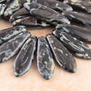 May include: A selection of dark, elongated beads with a marbled pattern of black, white, and pale green. The beads are arranged in a circular pattern, highlighting their unique shape and colour variations. Suitable for jewellery making.