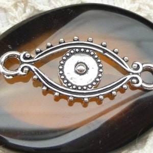 May include: A brown and black agate stone with a silver eye charm. The charm has a beaded border and a small silver bead in the center.