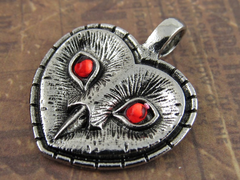 Peut inclure: Pendentif en forme de c&oelig;ur en argent avec un motif de visage de hibou. Le hibou a des yeux rouges et un bec d&eacute;taill&eacute;.