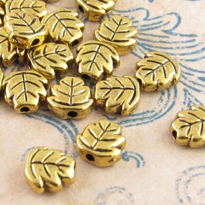 May include: A collection of small, gold-colored metal beads shaped like leaves. Each bead features a raised, embossed vein pattern on the surface and a single hole drilled through the center for stringing, creating a uniform, metallic appearance.