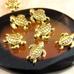 May include: Gold-colored turtle charms are scattered on a dark brown and tan oval-shaped stone. The charms have detailed shell patterns and are arranged in a visually appealing manner, suggesting jewelry components.