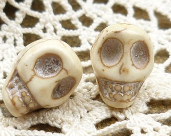 May include: Two white skull beads with carved details, including eye sockets and teeth. The beads are on a white crocheted surface.