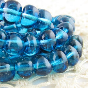 May include: A strand of teal blue glass beads. The beads are round and have a slightly irregular shape. They are strung on a clear thread.