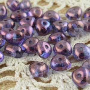 May include: A group of iridescent purple glass beads with a teardrop shape. The beads have a shiny, reflective surface and are arranged on a white crocheted surface.
