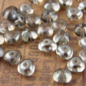 May include: A group of clear glass beads with a smoky gray tint. The beads are shaped like a teardrop with a flat bottom and a small hole in the center.