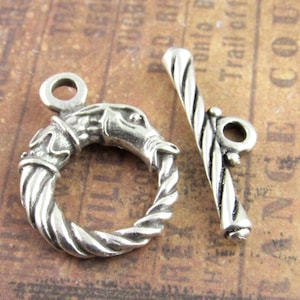 Può includere: Una chiusura a levetta argentata con un design dettagliato. La chiusura presenta un anello con un motivo a corda intrecciata e una testa di animale scolpita, insieme a una barra abbinata. La barra ha un design intrecciato e un piccolo anello.