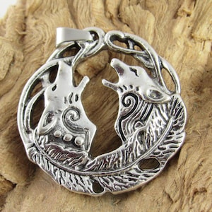 May include: Silver pendant depicting two howling wolves in a circular design. The pendant has detailed patterns and is displayed on a textured, light brown wooden surface. The pendant is likely made of a metal alloy.