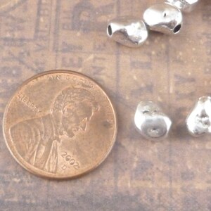 May include: A close-up of a collection of silver metal beads. The beads are irregular in shape and have a hammered texture. The beads are scattered on a light brown surface. A US penny coin is also visible in the image.