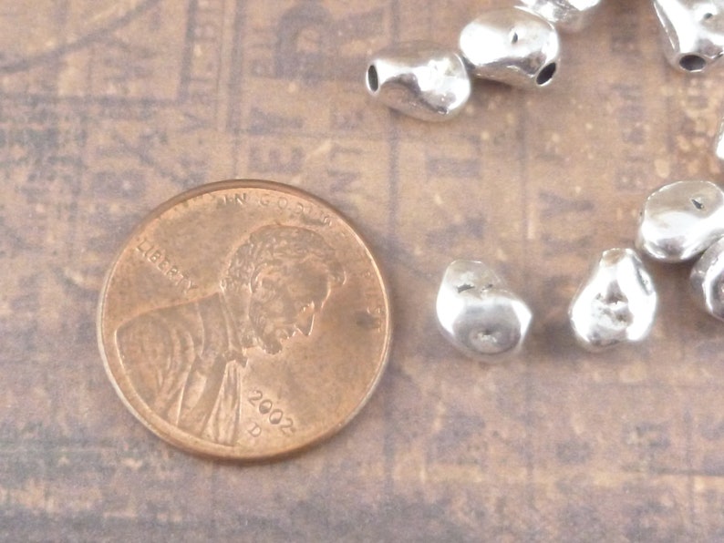 May include: A close-up of a collection of silver metal beads. The beads are irregular in shape and have a hammered texture. The beads are scattered on a light brown surface. A US penny coin is also visible in the image.