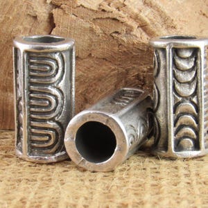 Puede incluir: Tres cuentas de metal plateado con intrincados diseños en relieve. Dos cuentas están de pie, mostrando un patrón repetitivo, mientras que la tercera yace horizontalmente, revelando un agujero central. Las cuentas cilíndricas tienen un aspecto envejecido.