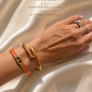 May include: Two gold-toned bracelets, one featuring a bright orange enamel inlay and a central oval link, the other shaped like a bent nail. Text on the image reads: 18K GOLD, WATERPROOF, TARNISH RESISTANT, HYPOALLERGENIC. A diamond ring is also visible.