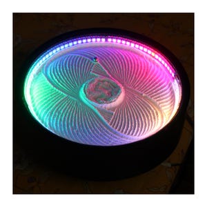 Könnte beinhalten: Ein runder kinetischer Sandkunsttisch mit schwarzem Rahmen und einem internen LED-Lichtstreifen, der die weiße Sandoberfläche in leuchtenden Rosa-, Blau- und Grüntönen beleuchtet. Eine kleine Metallkugel liegt in den komplexen Sandmustern.