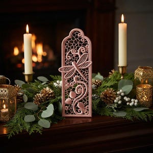 May include: A vertical, arched, dusty pink lace ornament featuring a dragonfly motif with intricate openwork patterns. The piece is displayed among green foliage, pinecones, white berries, and two tall cream-colored candles in gold holders.