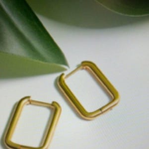May include: A pair of gold-toned hoop earrings featuring a rectangular shape with rounded corners. The earrings have a smooth, polished finish and a hinged post mechanism for secure closure, presented as two distinct pieces.