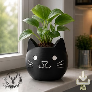 Puede incluir: Una maceta negra mate con forma de gato que presenta detalles blancos grabados para los ojos, la nariz y los bigotes. El recipiente redondo contiene una planta de hojas verdes vibrantes en forma de corazón plantada en tierra oscura.