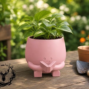 May include: A matte pink ceramic planter shaped like a small character holding an open book. The planter features a smiling face, two arms, and two feet. A green leafy plant is potted inside the container, which sits on a wooden surface.