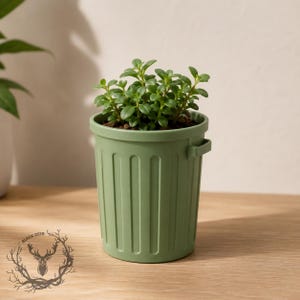 May include: A small green plant with rounded leaves potted in a sage green plastic container shaped like a miniature trash bin. The container features vertical ribbed detailing along the sides and a small handle on the rim. The plant is centered.