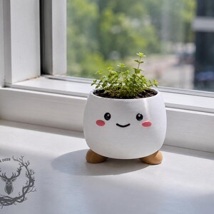 Pode incluir: Um vaso branco e arredondado com acabamento liso, apresentando um rosto impresso com olhos pretos, um pequeno sorriso e bochechas cor-de-rosa. O vaso assenta sobre três pequenos pés arredondados castanho-claros e contém uma pequena planta verde.