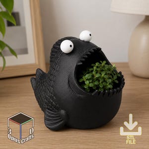 May include: A black, fish-shaped planter with a wide, open mouth containing a small green plant. The fish has two large, white, round eyes with black pupils positioned above its mouth. The text 3D Opus Crafts and an STL file icon are visible.