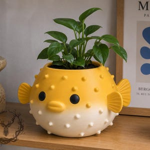 May include: A yellow and white pufferfish-shaped ceramic planter featuring a gradient color design, small raised spikes, two black circular eyes, and a small mouth, containing a green leafy plant with multiple stems and oval-shaped leaves.