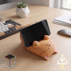 May include: An orange tabby cat-shaped phone stand made of matte material, featuring a textured striped pattern, small ears, and raised paws that hold a black smartphone in a horizontal position. The base includes the text 3D Opus Crafts.