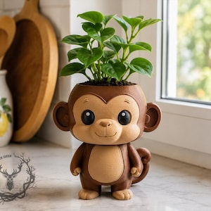 May include: A brown and tan monkey-shaped planter featuring a smooth, matte finish, large round ears, and a small tail. The head serves as the container for a small green plant with rounded leaves. A logo with a deer and the text BLACK DEER is visible.