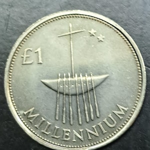 May include: A silver coin featuring a stylized boat with a mast and two stars. The coin has the text "£1" and "MILLENNIUM" around the edge. The coin has a textured edge and a smooth, reflective surface.