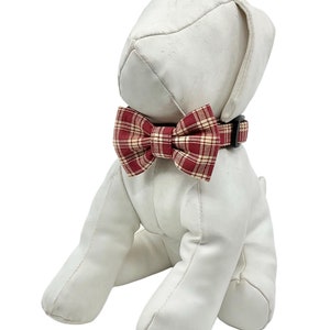 May include: White stuffed dog wearing a red and tan plaid bow tie. The bow tie is tied around the dog's neck with a black adjustable collar.