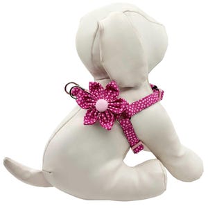 May include: A white stuffed dog wearing a pink polka-dot harness with a large flower accent. The flower and harness are a vibrant pink with white polka dots. The dog is sitting with its back to the viewer.