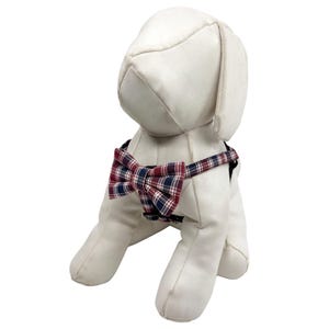 May include: A white stuffed dog wearing a plaid bow tie and harness. The bow tie features a pattern of red, navy, and white. The dog is posed sitting down, facing slightly to the side.