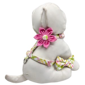 May include: A white plush dog wearing a pink and green plaid harness with a matching bow tie. The harness features a large pink flower accent on the back and a metal ring for leash attachment.