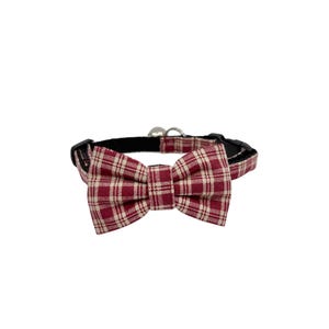 May include: A cat collar with a burgundy and cream plaid bow tie. The collar is black with a silver bell and a plastic buckle. The bow tie is centered on the collar.