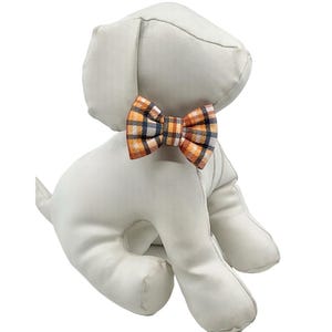 May include: A stuffed dog toy in a sitting position, made of a soft, light gray fabric. It features a plaid bow tie in orange, navy, and white. The toy is designed to resemble a puppy.
