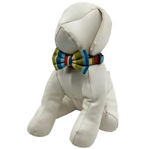 May include: A white stuffed dog wearing a colorful striped bow tie and collar. The bow tie features stripes of blue, green, red, and yellow. The collar has a matching striped pattern. The dog is sitting.