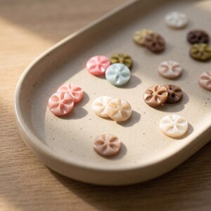 Puede incluir: Una colección de botones pequeños y redondos en varios colores pastel y tonos tierra, cada uno con una hendidura central en forma de flor con seis pétalos. Los botones están esparcidos sobre una bandeja de cerámica beige claro, ovalada y lisa.