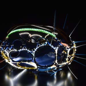 May include: A spherical structure composed of interconnected blue circular rings and dark blue fabric panels, illuminated by integrated white and green LED light strips. The object features protruding white plastic cable ties throughout.