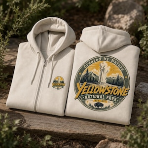 May include: A cream-colored zip-up hoodie featuring a small Yellowstone National Park logo on the left chest and a large circular graphic on the back depicting a geyser, mountains, and a bison with the text Created by Nature Yellowstone National Park EST 1872.
