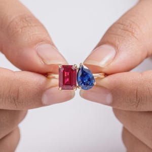 May include: A gold ring featuring two prominent gemstones set side by side. One stone is a vibrant red emerald-cut ruby, and the other is a deep blue pear-cut sapphire. The stones are held in place by small metal prongs on a thin gold band.