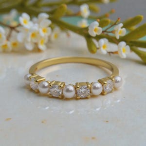 Puede incluir: Un anillo de oro con un patrón alterno de perlas blancas redondas y piedras transparentes facetadas engastadas en una fila a lo largo de la mitad superior de la banda. La banda de metal es fina y pulida, mostrando un tono oro amarillo brillante.