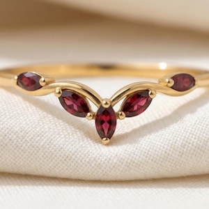 Könnte beinhalten: Ein goldener V-förmiger Ring mit fünf tiefroten Granat-Edelsteinen im Marquise-Schliff, die symmetrisch entlang der Ringschiene angeordnet sind. Die polierte Goldschiene läuft spitz zu, wobei die Steine sicher in Goldfassungen sitzen.