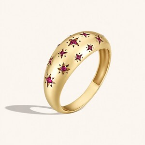 May include: A gold dome ring featuring a polished finish and multiple star-shaped engravings. Each star cutout is centered with a small, round, vibrant pink gemstone, creating a celestial pattern across the curved surface of the band.