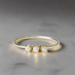 May include: A thin gold band ring featuring three small, round, white pearls set horizontally in a row. The pearls are secured by individual prongs along the centre of the band, creating a delicate and balanced design on the polished gold metal.
